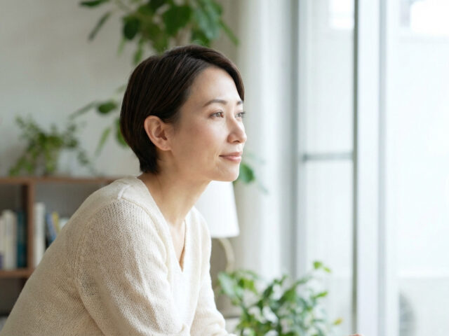 Femme aux cheveux courts, pull clair, regardant pensivement par la fenêtre dans un intérieur lumineux avec plantes et livres.