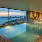 Vue d'une piscine intérieure d'un spa de luxe avec une baie vitrée donnant sur la mer et des rochers au crépuscule. Un couple se repose.