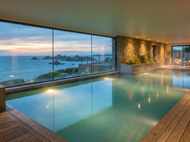 Vue d'une piscine intérieure d'un spa de luxe avec une baie vitrée donnant sur la mer et des rochers au crépuscule. Un couple se repose.